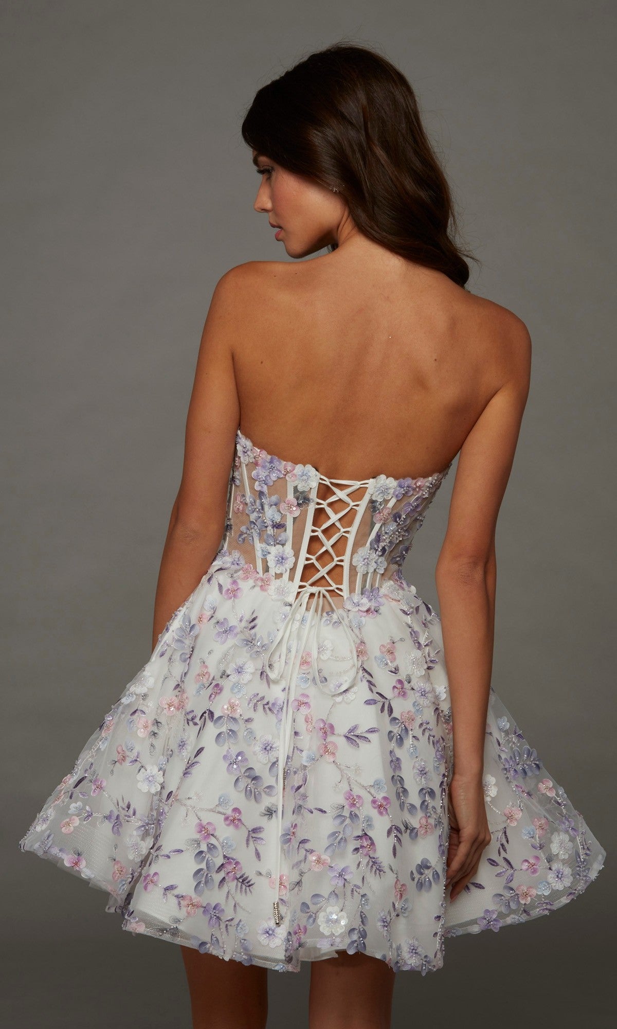 A woman stands with her back turned, wearing the strapless fit-and-flare Alyce 30086 short homecoming dress, which showcases a lace-up back and pastel embroidery with delicate 3D floral accents, set against a plain gray background.